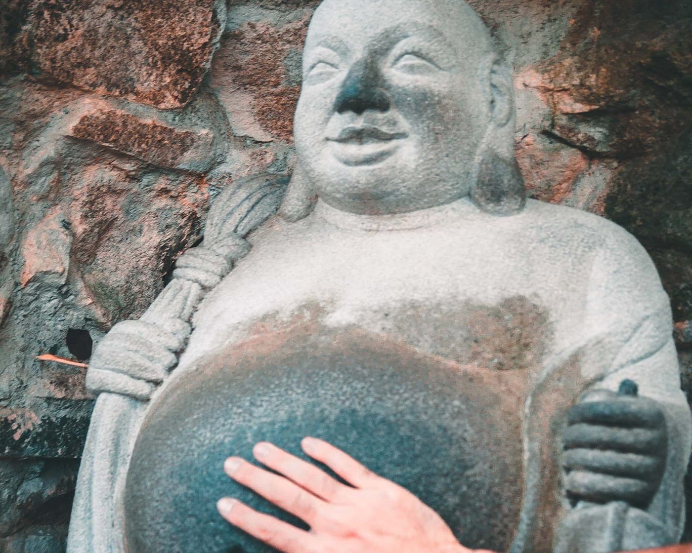 A hand resting on the belly of a carved stone Buddha set into a rock wall.